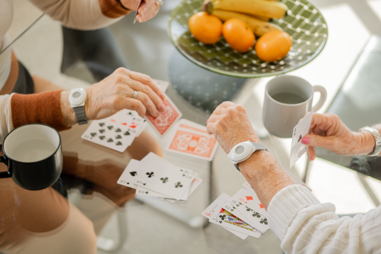 Two pairs of elderly hands wearing Freedom Medical Alarm wrist pendants play cards.