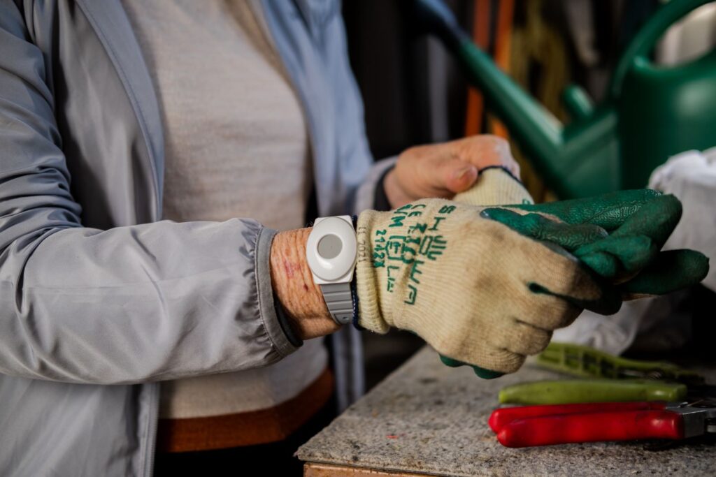 An elderly woman wearing a medical alarm wrist pendant takes off gardening gloves.
