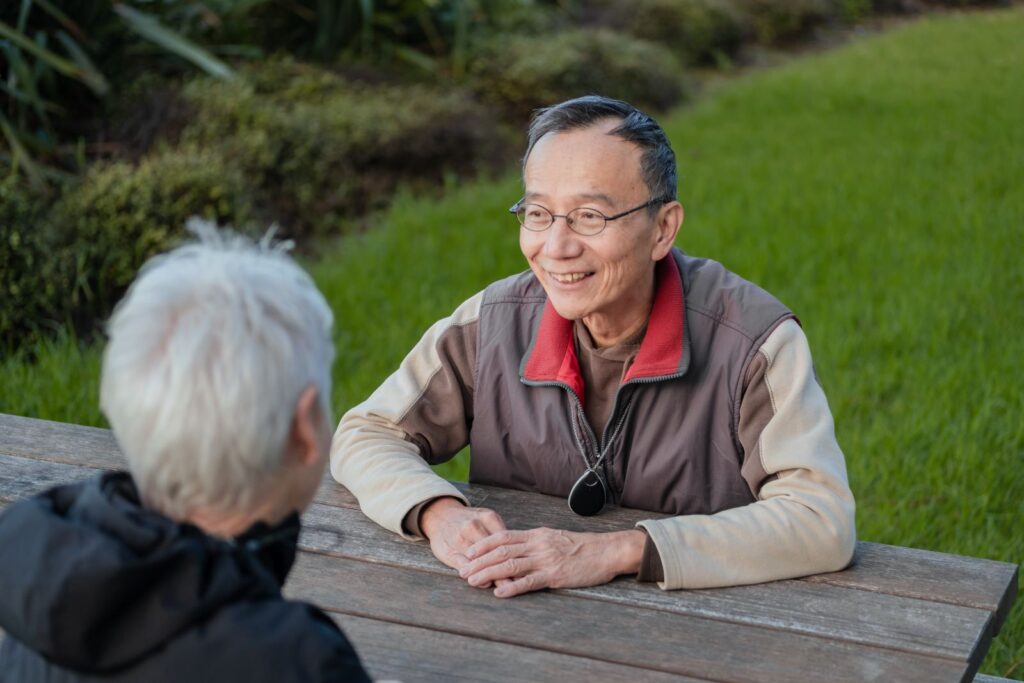 A elderly man wearing a Freedom Medical Alarm sits at a park bench with a woman.