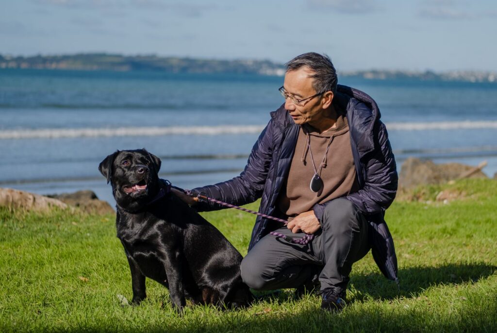 An elderly man kneels by his dog beside the beach.